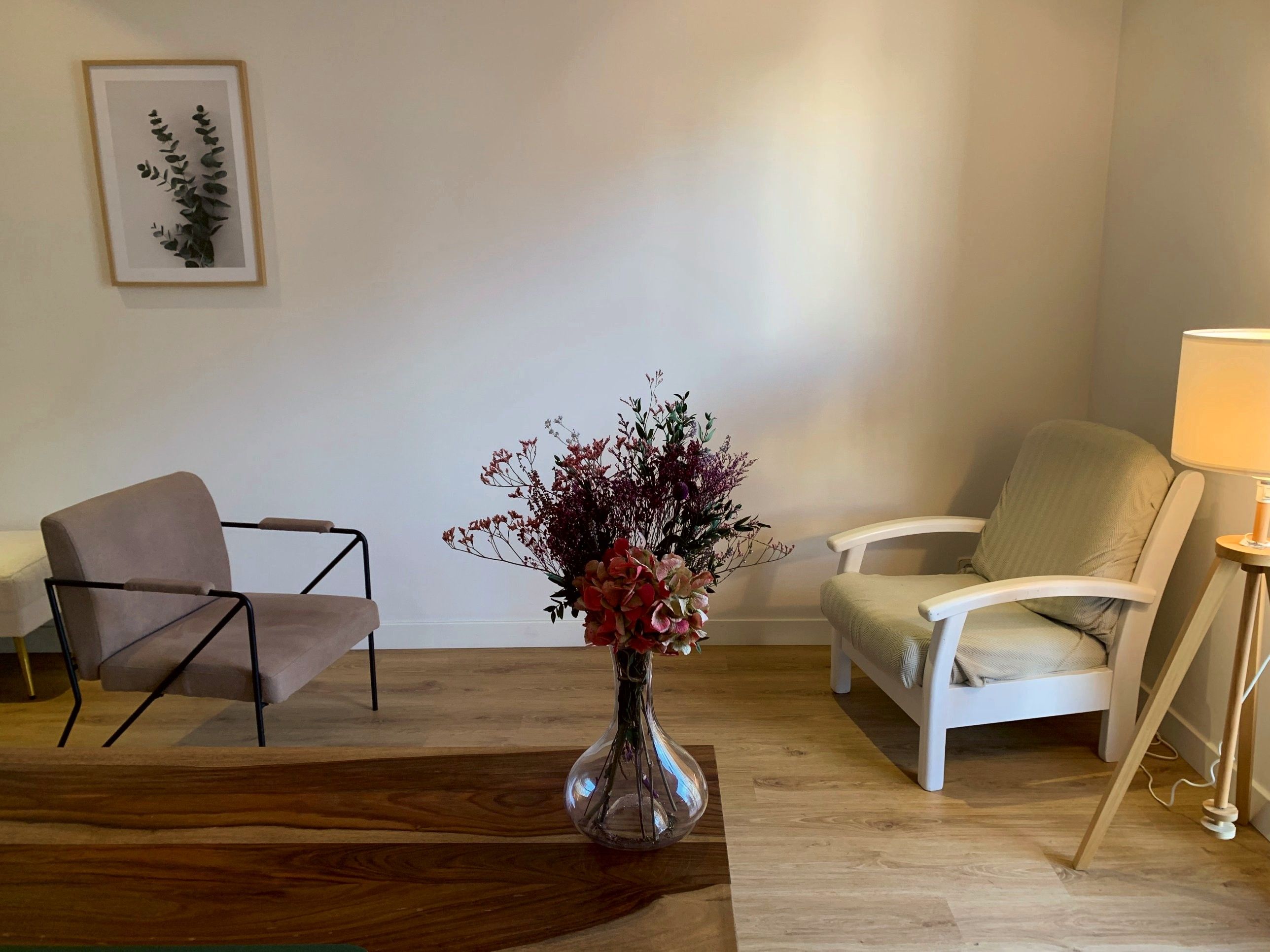 Sala de la consulta con dos butacas, un cuadro de eucalipto, una lámpara de pie y flores secas sobre la mesa