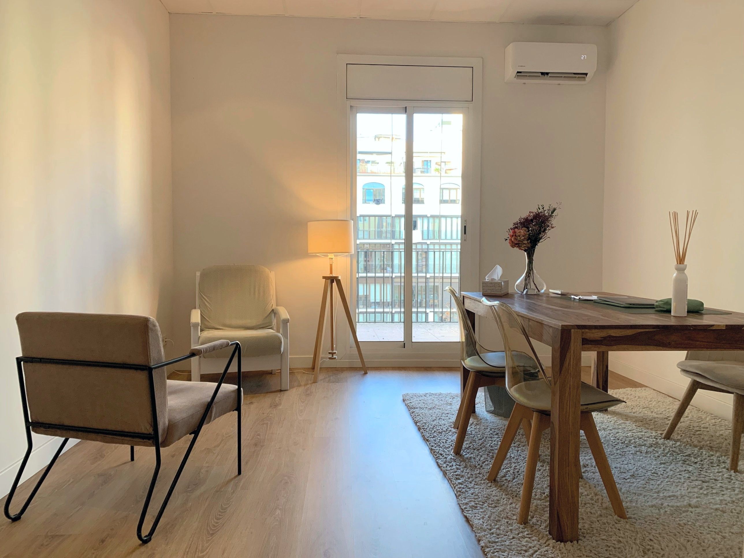 Vista general de la consulta: dos butacas y una lámpara junto al balcón, y una mesa de madera con sillas sobre una alfombra clara